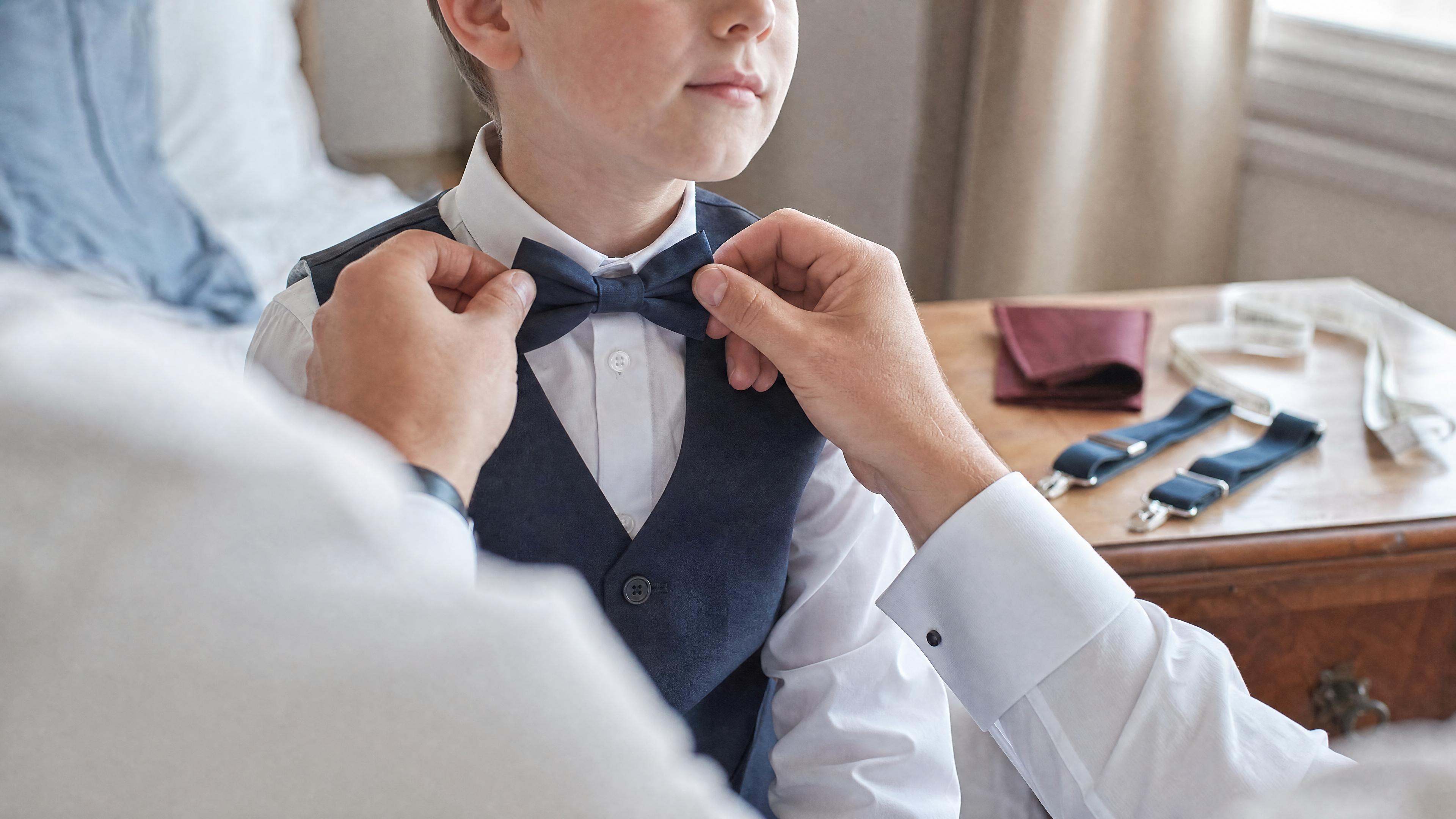 Un adulte ajuste un nœud papillon sur un enfant en chemise blanche, avec un mètre ruban visible sur une commode, dans une chambre lumineuse avant une cérémonie Imagem